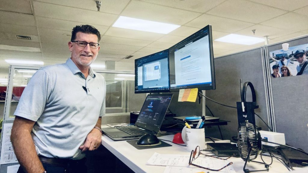 A smiling man with glasses and a striped polo shirt leans against an office cubicle desk equipped with dual monitors and a laptop.