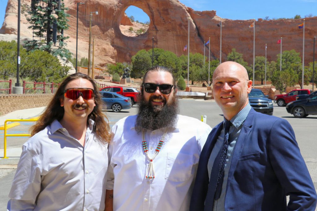 Three men stand smiling together outdoors in a sunny parking lot, with the red sandstone Window Rock natural arch formation visible in the background.