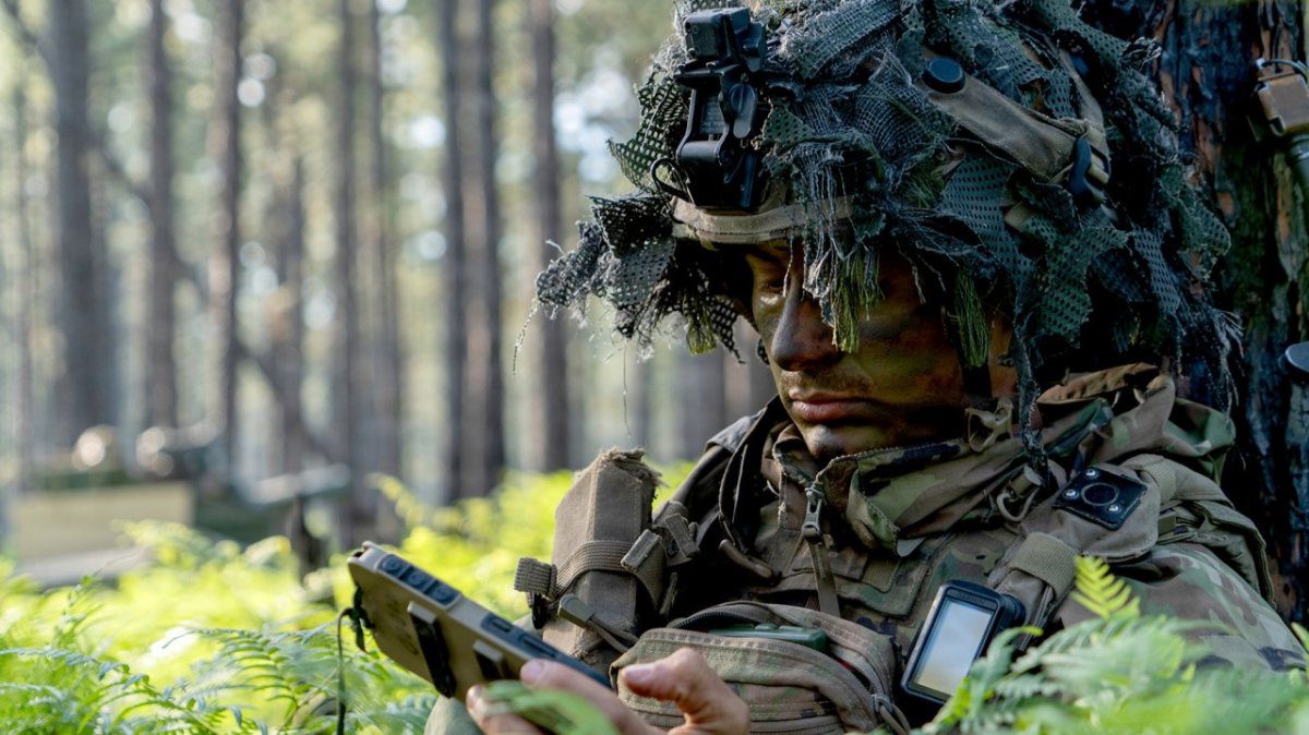 A soldier with camouflage face paint and a foliage-covered helmet looks down at a rugged handheld mobile device while leaning against a tree trunk in a dense forest.