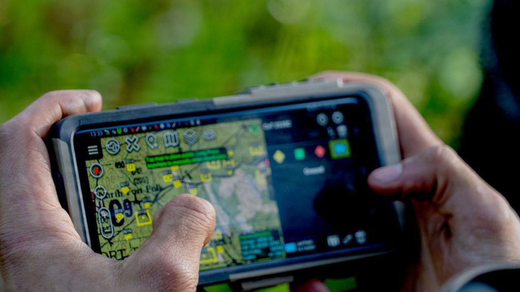 A close-up of hands holding a rugged mobile screen showing an active tactical topographical map application filled with waypoint icons, grids, and operation menus.