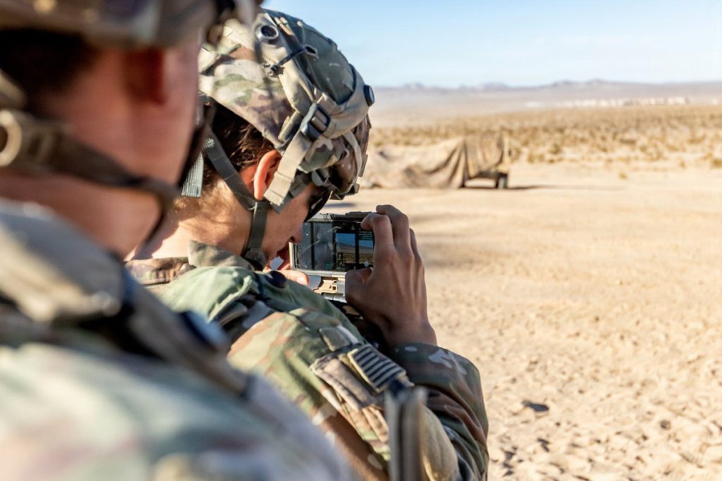 An over-the-shoulder view of a soldier in a desert environment holding up a ruggedized smartphone device horizontally to capture geospatial data or coordinate field operations.