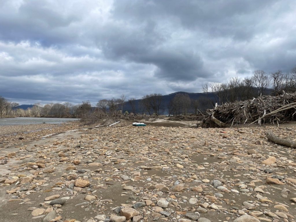 A rocky shoreline alongside a wide river leading to a massive pile of accumulated woody debris against distant mountain ridges under a cloudy sky.