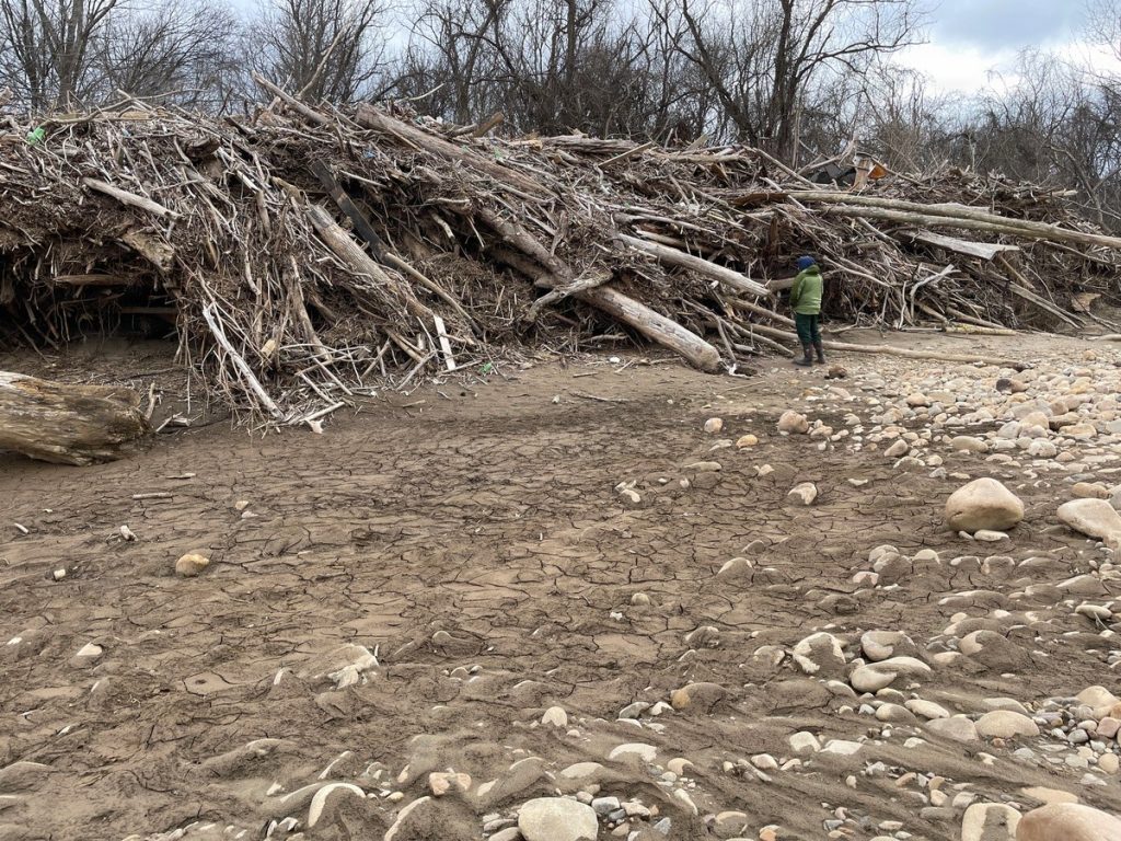 A lone inspector in a green jacket stands beside a towering pile of logs and vegetative debris piled on a cracked, dry river bed.