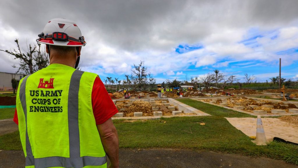 A USACE safety inspector seen from behind in a high-visibility yellow vest and hard hat looks out over an active foundation construction area.