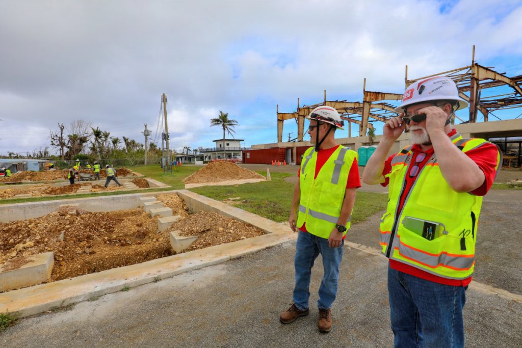 Two safety inspectors wearing white hard hats and high-visibility yellow vests observe concrete foundation trenches and structural work at a building site.