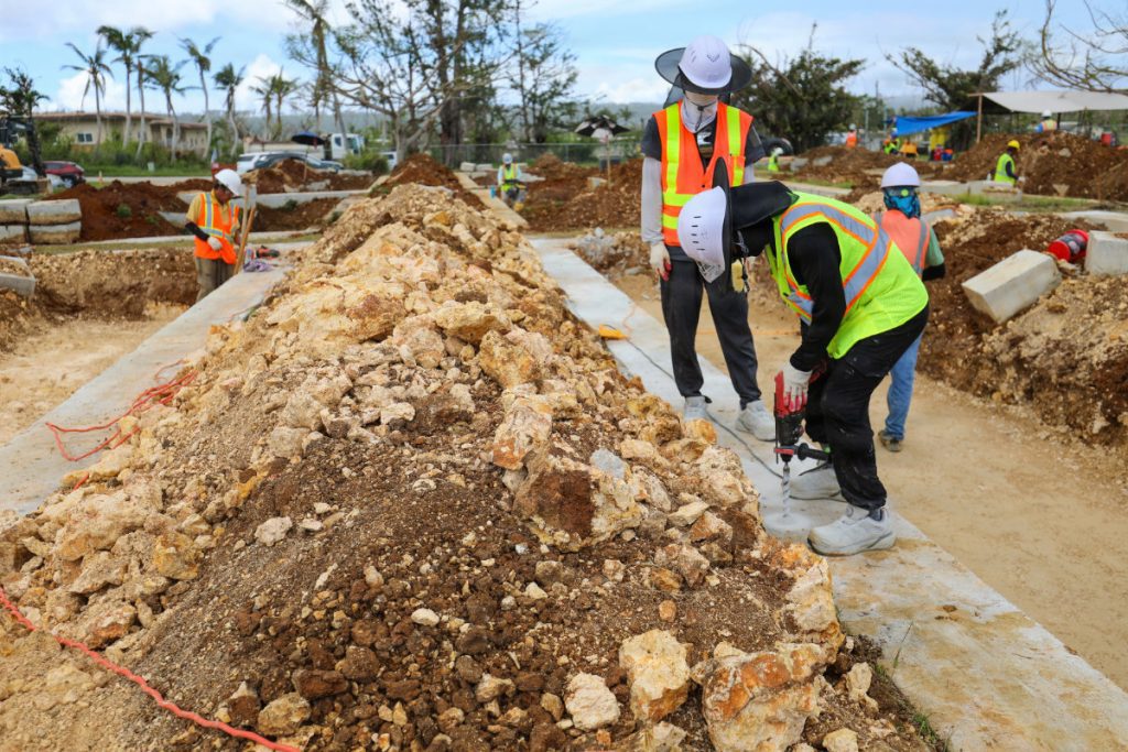 Construction workers in protective gear and hard hats use power drills into concrete footings beside large piles of earth and gravel.