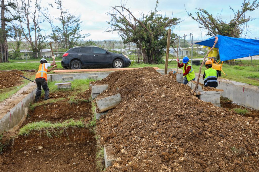 Construction crew members use pickaxes and shovels to dig out foundation trenches near earth piles and temporary shelters.