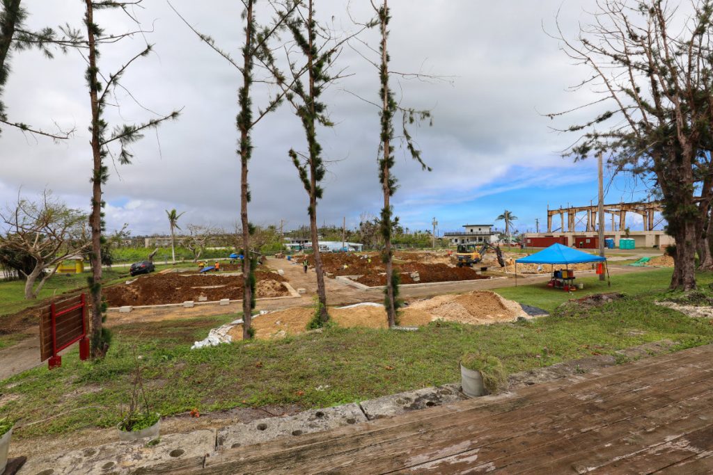 A wide view of an outdoor school construction site with dirt foundations, heavy machinery, tall pine trees, and a blue canopy shelter under a cloudy sky.