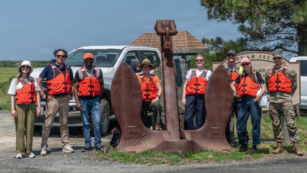 A group of military and international embassy officials wearing orange life jackets pose for a photo behind a massive, historic rusted ship anchor next to a white pickup truck.