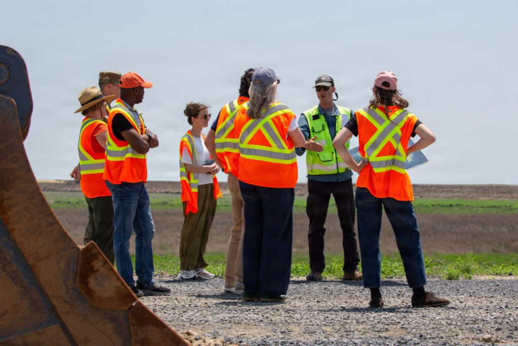 A group of international officials and engineers wearing bright orange and yellow safety vests stand in a circle on a gravel path, listening to a field briefing next to a large excavator bucket.