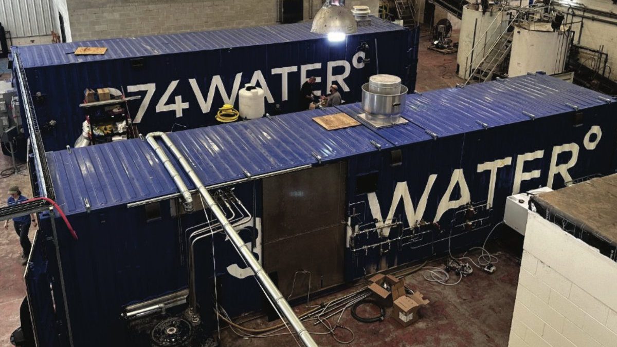 An overhead view of two large blue shipping containers with white "374WATER" branding housed inside an industrial facility with piping and equipment.
