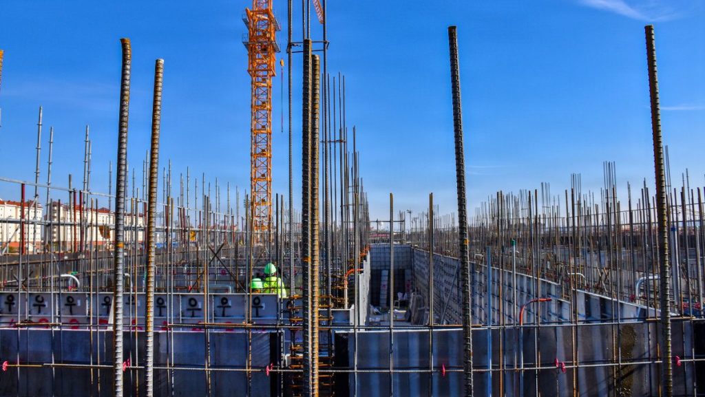 Looking through a construction framework at a foundation site filled with rows of metal reinforcement rebar columns and scaffolding under a clear blue sky, with a yellow tower crane standing in the background.