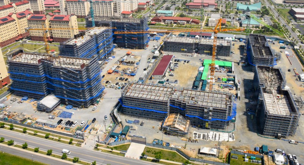 An aerial bird's-eye view of a large military housing complex construction site, showing several multi-story buildings wrapped in blue safety mesh and scaffolding with multiple tall yellow cranes operating.