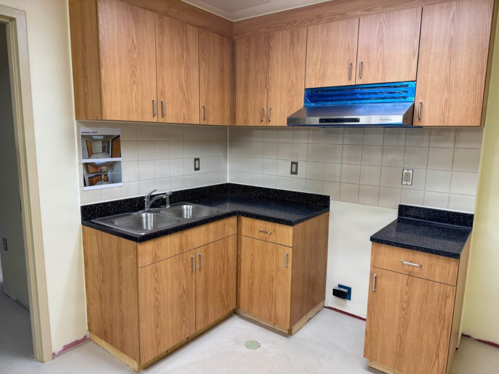 The interior of a newly built barracks apartment kitchen featuring light wood cabinetry, dark speckled stone countertops, a double stainless steel sink, and a modern range hood.