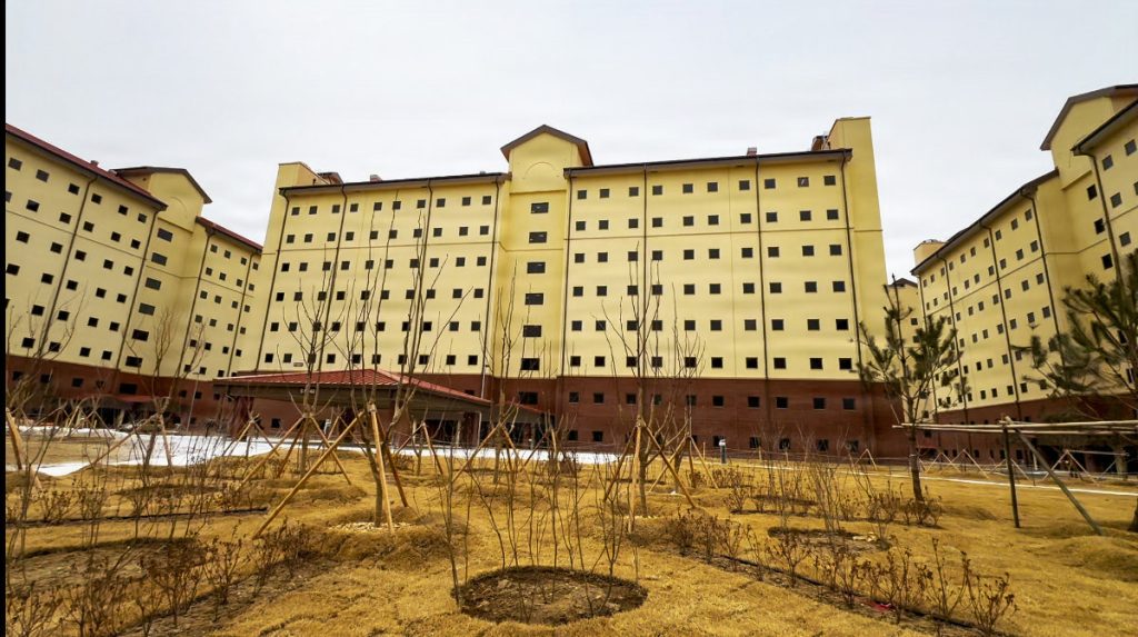 An exterior view of newly finished multi-story yellow and brown military barracks buildings under a cloudy sky, featuring landscaped grounds with small trees supported by wooden stakes in the foreground.