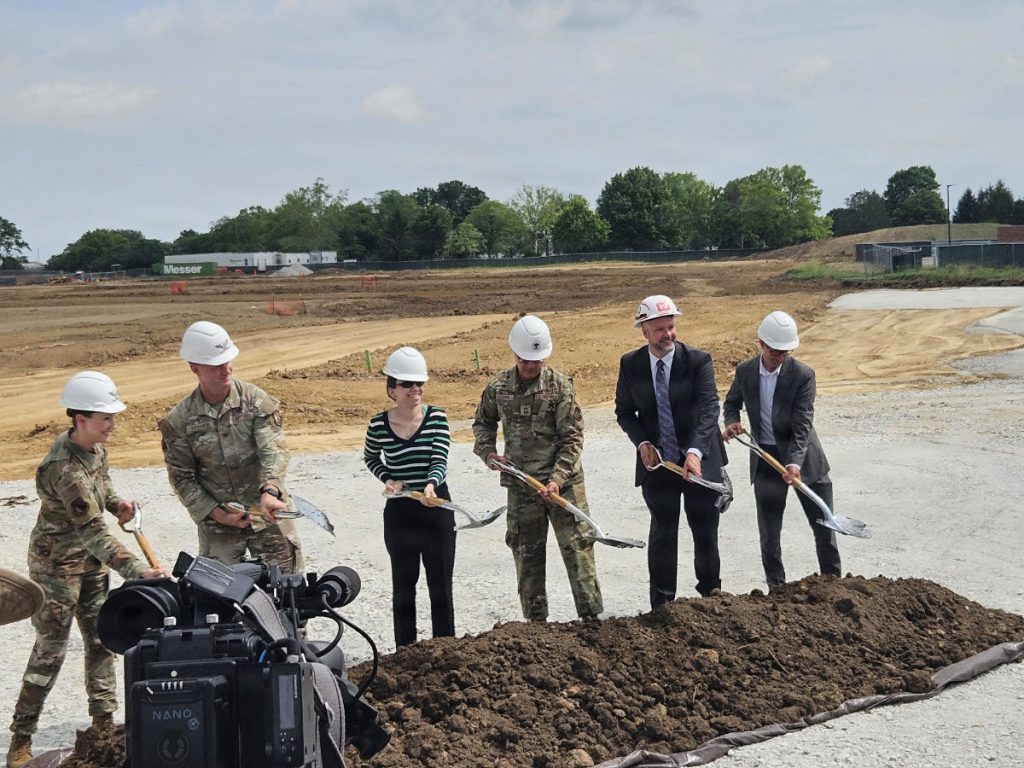 Military personnel in camouflage uniforms and civilians wearing hard hats stand in a row, using shovels to break ground at a construction site with dirt fields behind them.