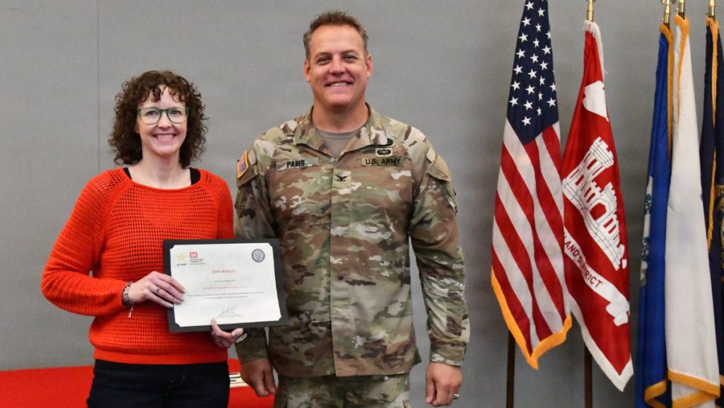 A woman in an orange sweater and glasses smiles while holding an award certificate beside a U.S. Army colonel in camouflage uniform in front of military flags.