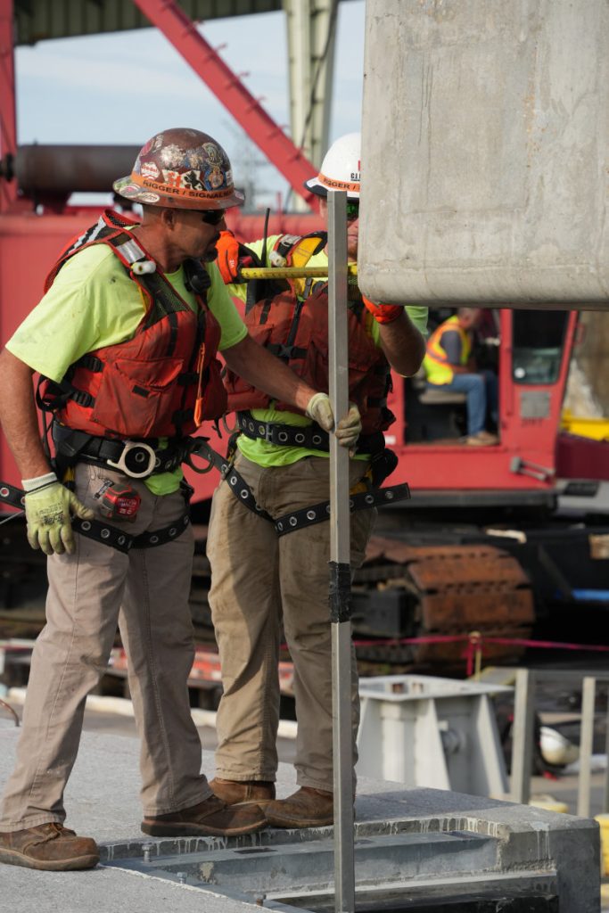 Two riggers wearing safety harnesses, life vests, and hard hats use a measurement tool to align a heavy concrete beam during a crane lift.