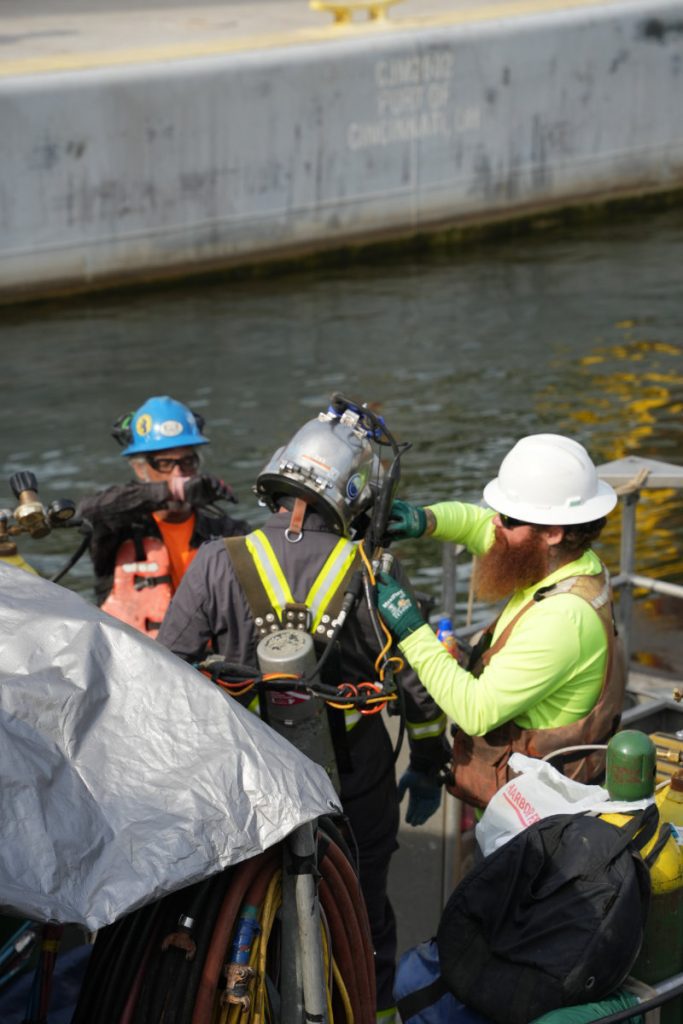 A commercial diver in full dive gear and helmet receives assistance from a surface support crew member on a boat deck near a concrete lock wall.