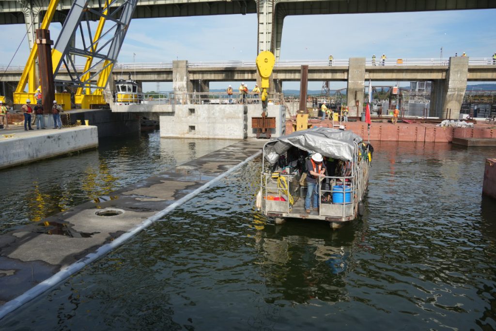 A technical workboat covered by a grey tarp navigates the river water within a concrete lock chamber canal during an active construction project.