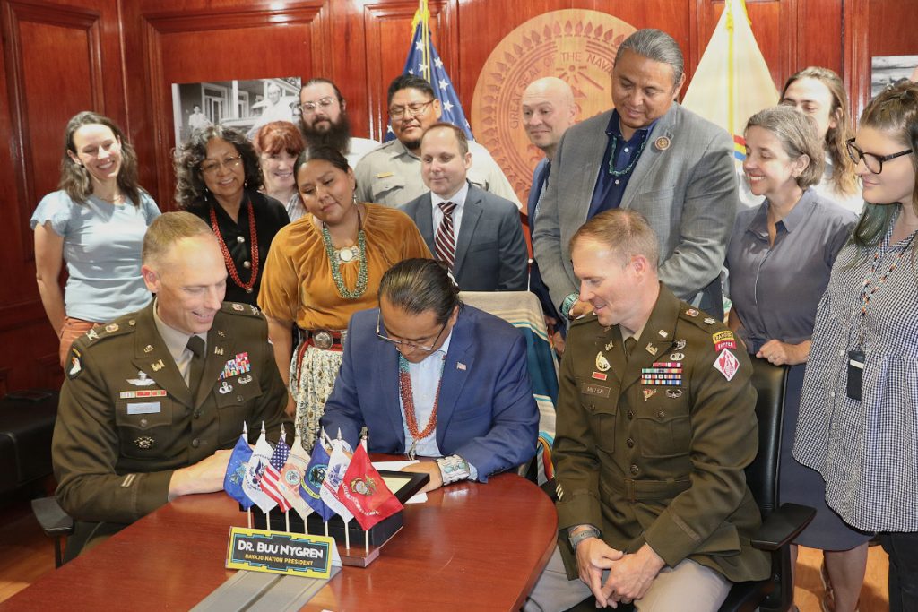 Navajo Nation President Dr. Buu Nygren sits at a wooden table signing an official document, flanked by two U.S. Army officers in dress uniforms and surrounded by tribal and federal officials.