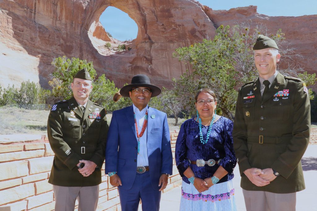 Two U.S. Army officers in service uniforms and two Navajo Nation leaders pose together outdoors, with the natural sandstone Window Rock arch framing the background.