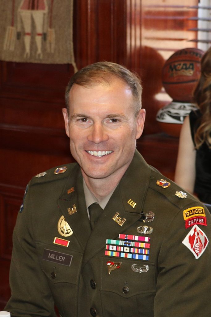 A portrait of a smiling U.S. Army officer, LTC Miller, in service dress uniform adorned with military medals, ribbons, and Ranger and Sapper tabs.