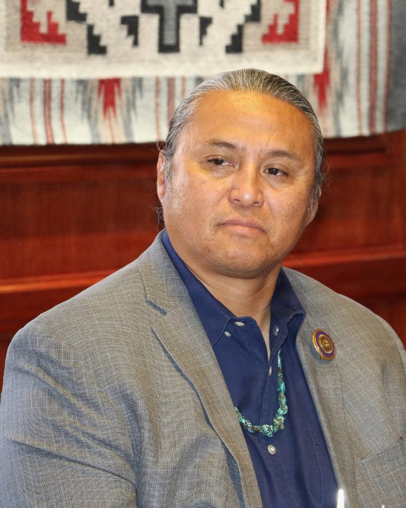 Close-up portrait of a Navajo Nation representative wearing a grey suit jacket, blue shirt, and a traditional turquoise bead necklace, sitting indoors.