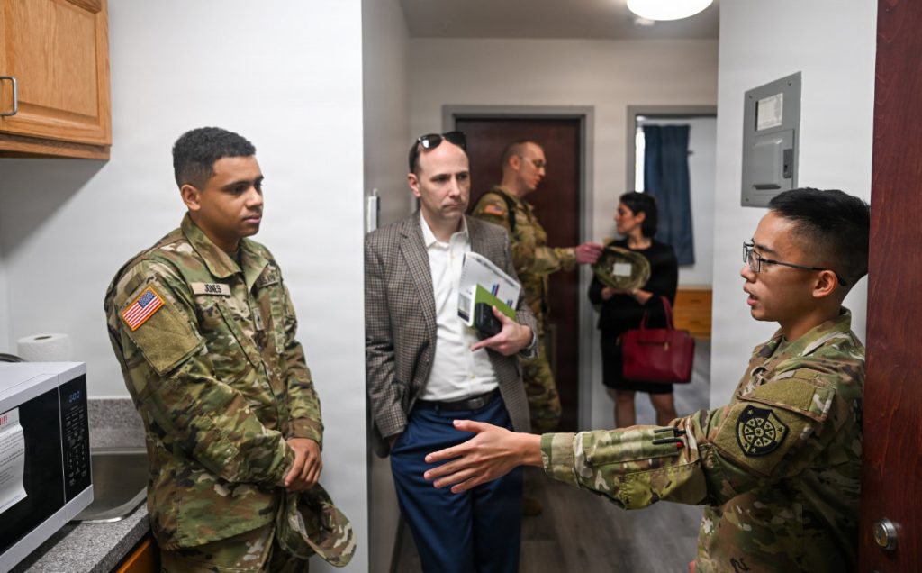 An Army soldier gestures and speaks to a civilian official and other military personnel standing inside a modernized barracks kitchen unit.