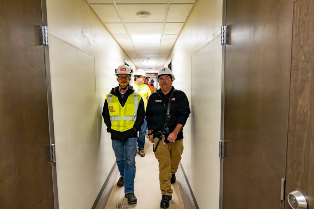 Safety officials and engineers wearing hard hats and safety gear walk down a brightly lit interior corridor of a barracks facility.