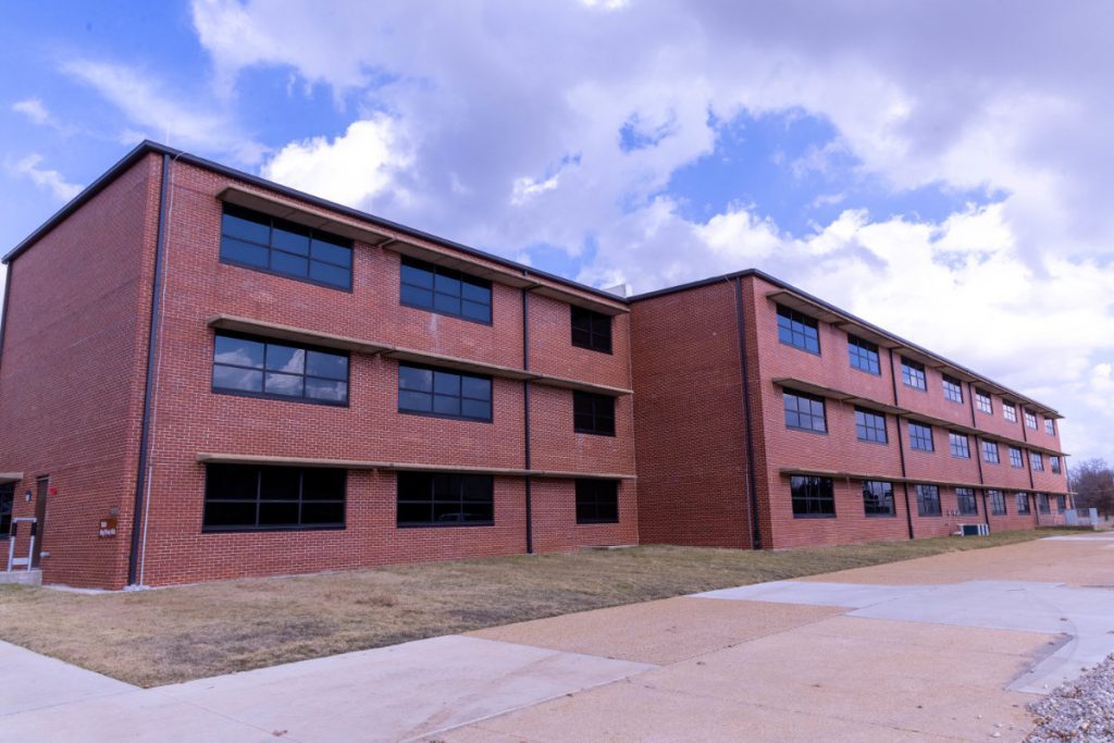 Exterior view of a multi-story red brick military barracks building with long rows of dark tinted windows under a cloudy blue sky.