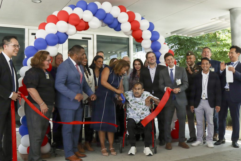 A group of military leaders, VA officials, and guests cheer under a red, white, and blue balloon arch while cutting a ceremonial ribbon for a veteran in a wheelchair.