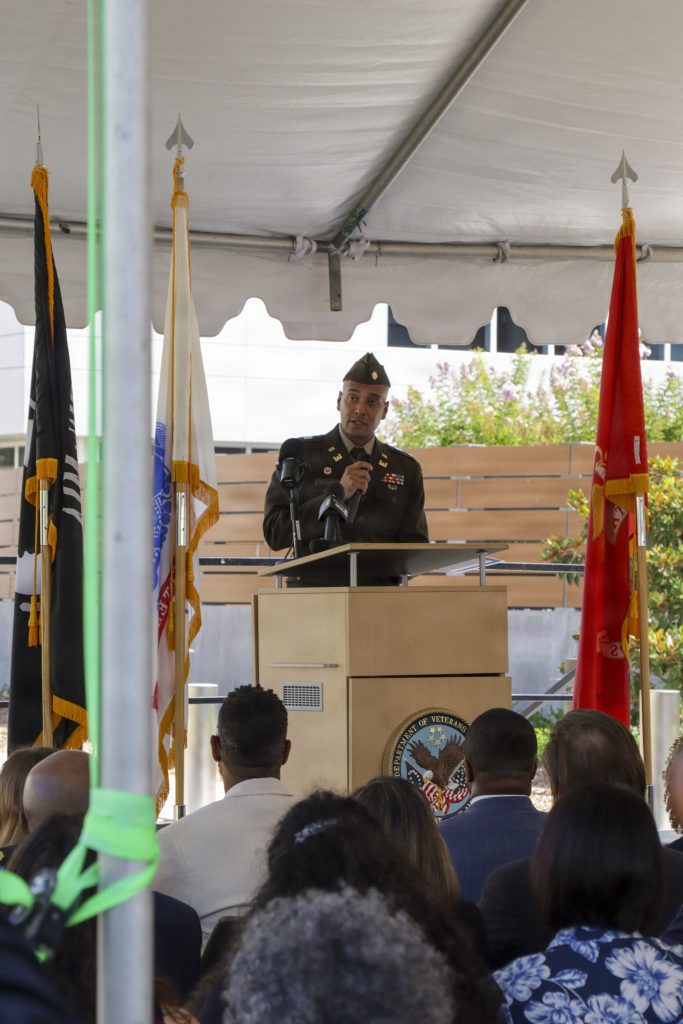 A U.S. Army officer in service uniform addresses an audience from a wooden lectern flanked by military flags under a white canopy tent.
