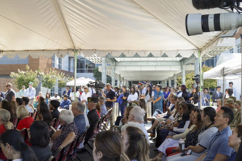 An audience of community members and medical staff sit in folding chairs under an outdoor tent listening to event speakers near the new facility entrance.