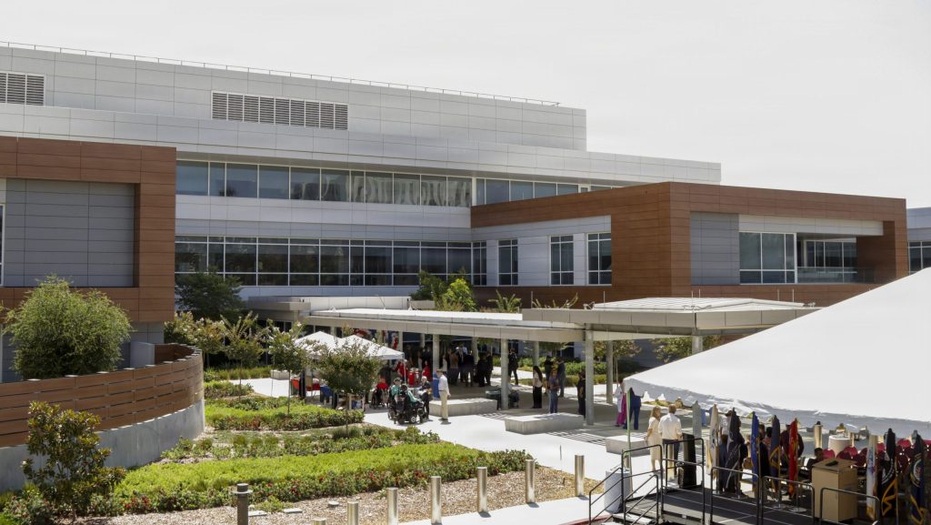 An exterior view of a modern multi-story medical facility with wood-paneled accents, courtyard landscaping, and event tents set up on the patio.