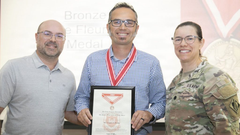 A man smiling and wearing a red medal ribbon around his neck holds an official certificate from the Army Engineer Association, flanked by a civilian man and a female U.S. Army officer.