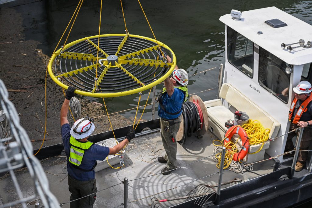 Workers in hard hats and safety vests stand on a utility workboat to steady a large, circular yellow steel ring lined with spiral black tubing suspended by a crane.