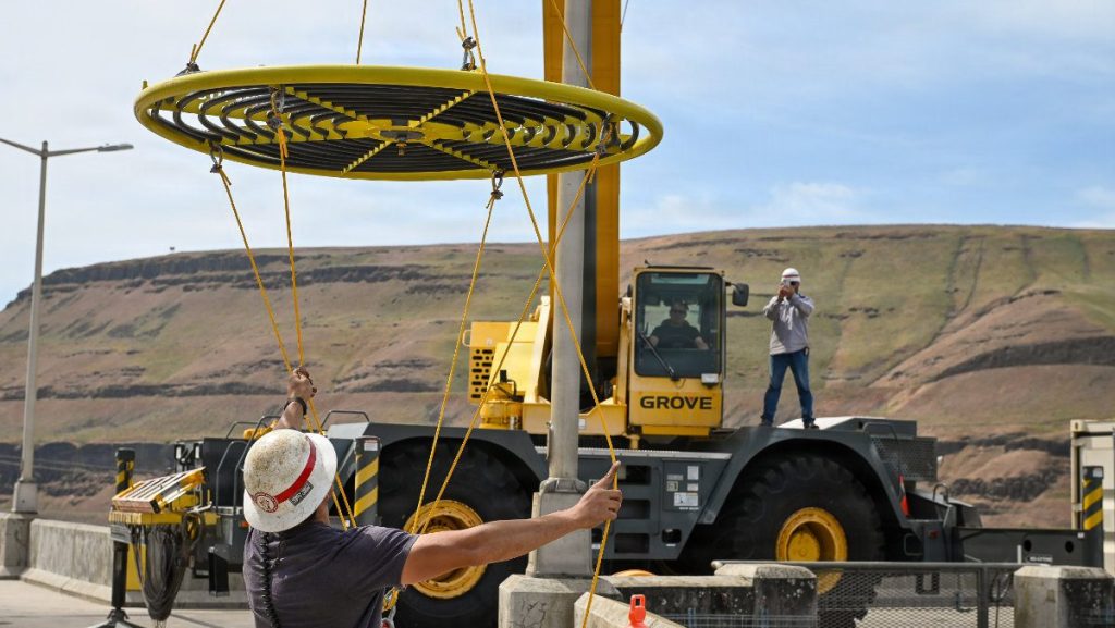 A ground worker uses guide lines to steady a large circular upwelling ring as a heavy-duty yellow Grove mobile crane lifts it high above a concrete deck.
