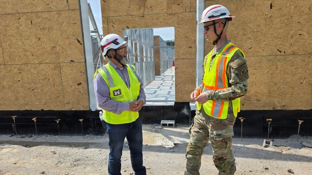 A civilian construction worker in a safety vest and hard hat converses with a U.S. Army officer in camouflage at an outdoor building site.