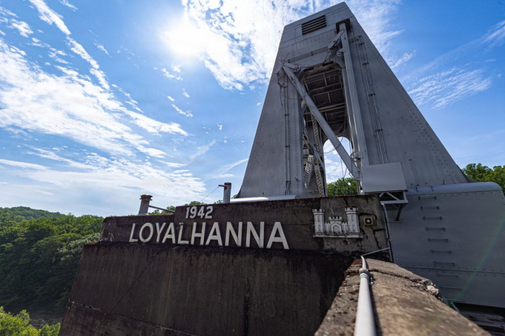 A weathered, rusted gantry crane sitting atop a dam structure beneath a bright blue sky with scattered clouds.