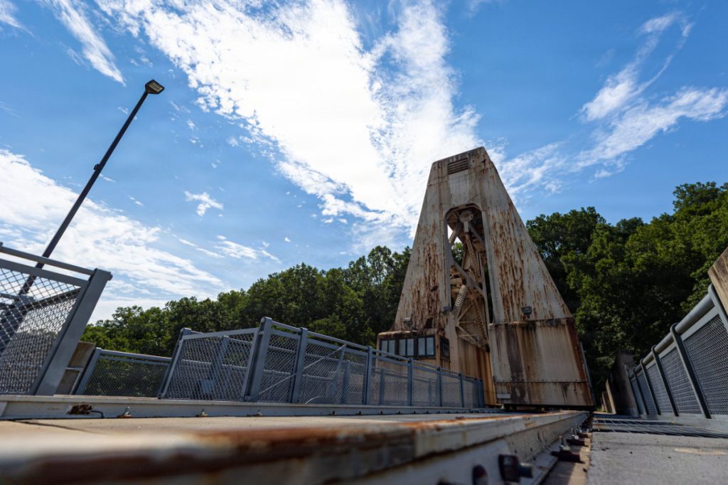 A weathered, rusted gantry crane sitting atop a dam structure beneath a bright blue sky with scattered clouds.