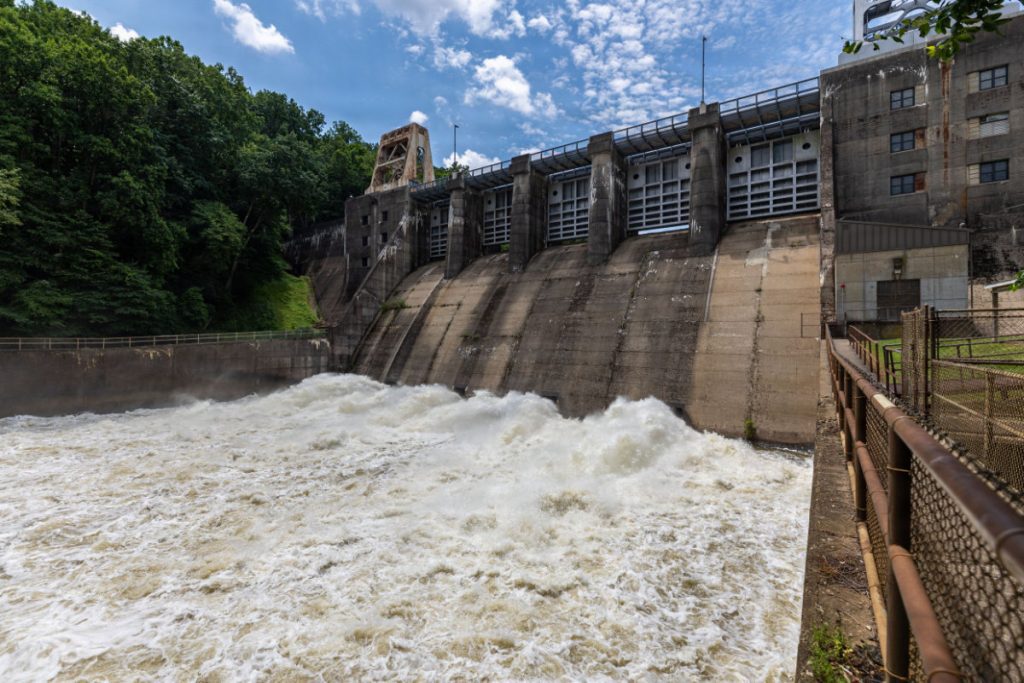 Rushing white water churns at the base of Loyalhanna Dam spillway gates with historic gantry cranes mounted along the top deck.