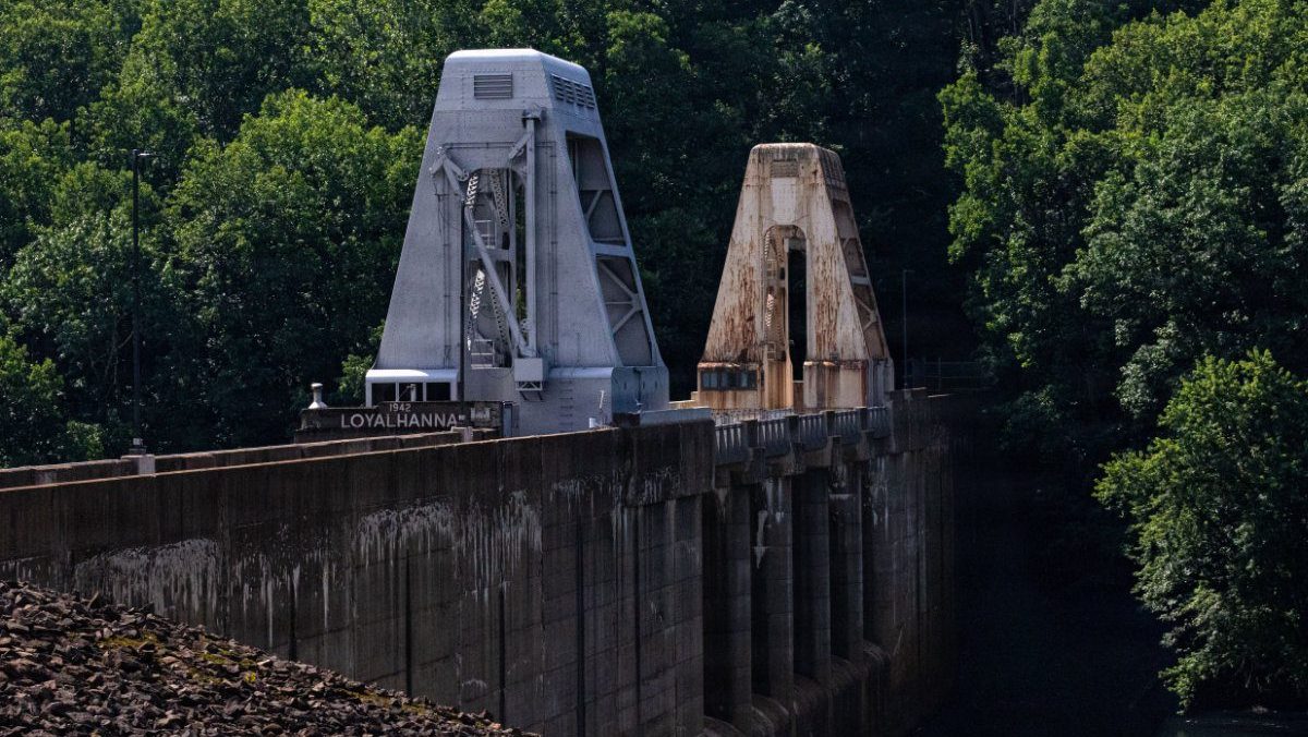 Two large dam gantry cranes—one freshly restored in grey and one weathered and rusted—stand side-by-side along the top of Loyalhanna Dam.