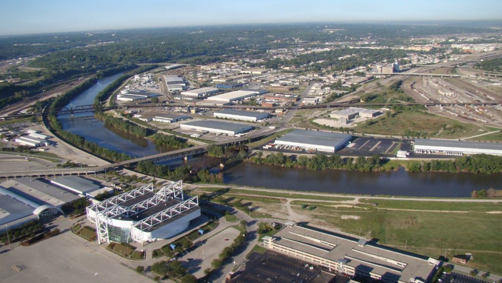 A wide high-altitude aerial photo showing the Kansas River winding past an arena facility, industrial warehouses, and rail yards.