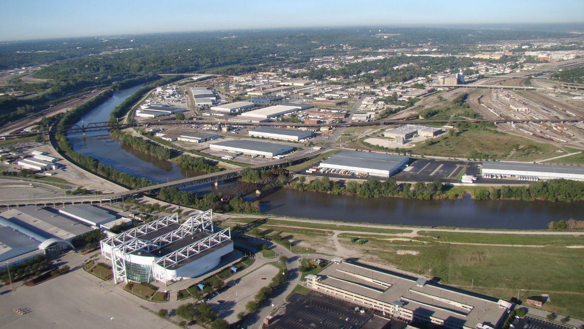 A wide high-altitude aerial photo showing the Kansas River winding past an arena facility, industrial warehouses, and rail yards.