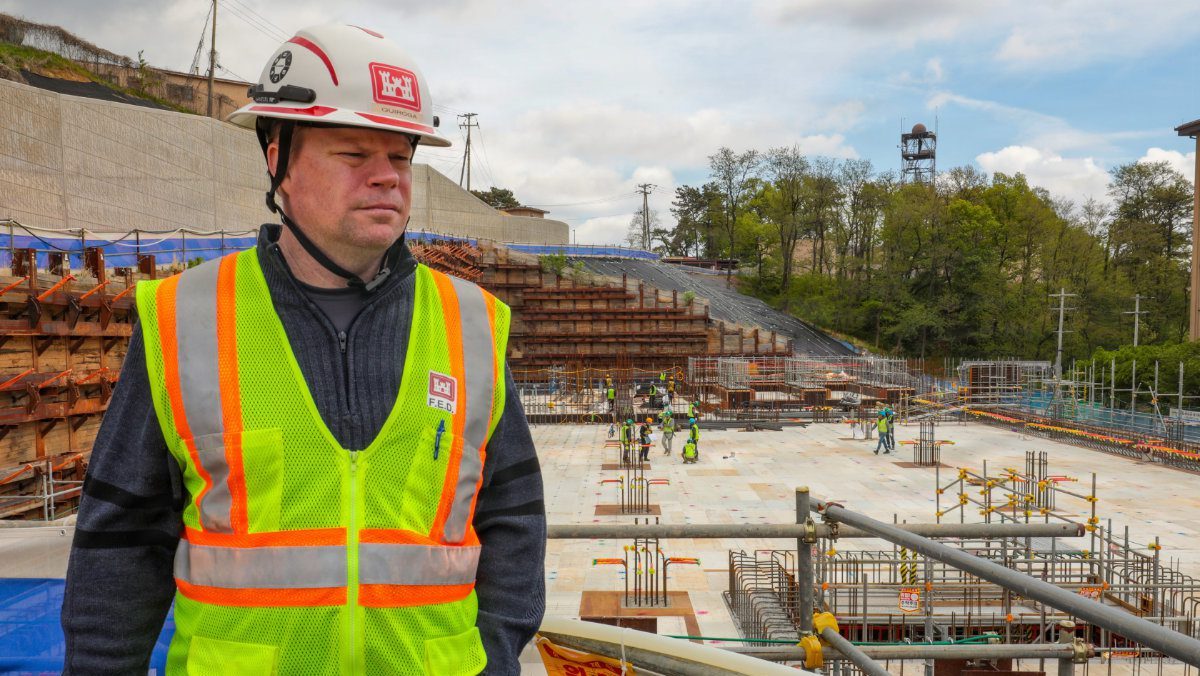 Brian Quiroga wearing a hard hat and high-visibility safety vest standing at an active construction site with retaining walls and foundation rebar in the background.