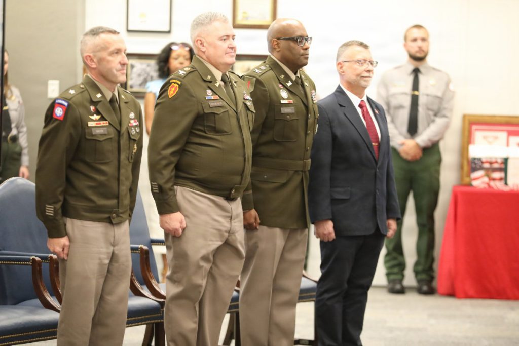 Four military and civilian leaders stand side-by-side at attention during a formal indoors ceremony with a park ranger standing in the background.