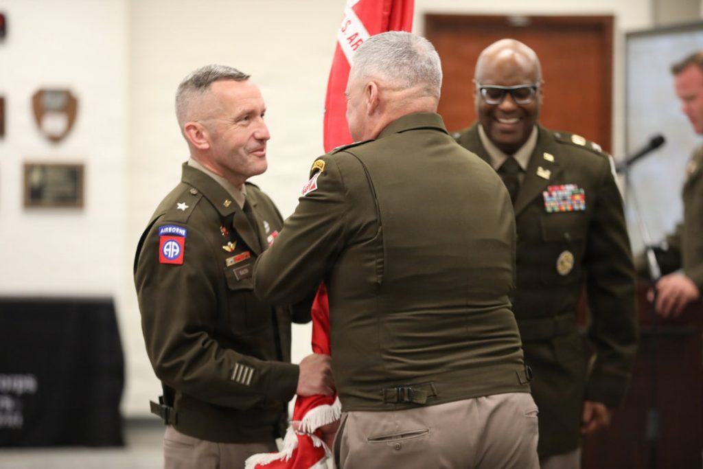 Two Army officers in service uniforms pass a red military flag back and forth while Col. Marlon Ringo smiles in the background.