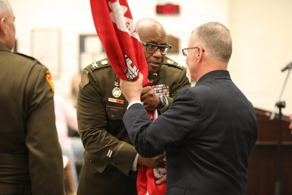 Col. Marlon Ringo in service uniform passes the red USACE organizational flag into the hands of a civilian leader during an official change of command ceremony.