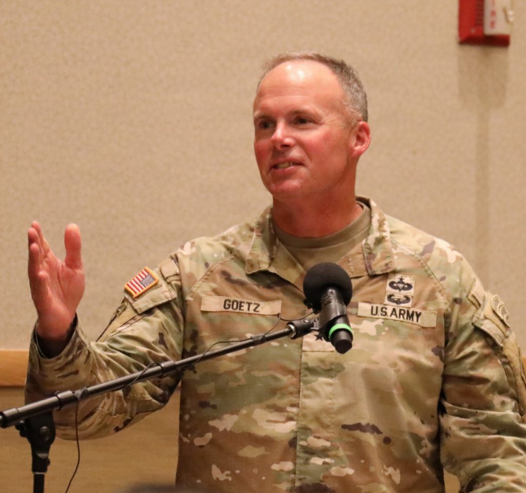 Brig. Gen. Kirk E. Gibbs in military uniform speaks into a microphone with his right hand raised while addressing attendees indoors.